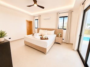 Ein modernes Schlafzimmer mit einem großen Bett, einem Deckenventilator, einem Nachttisch und Blick auf einen Swimmingpool.
