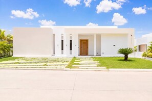 A modern two-story house with a flat roof, a front entrance with a wooden door, and a well-maintained lawn with a palm tree. - Modern Villa | Pool, Terrace & Sleeps up to 8 (Sosúa)