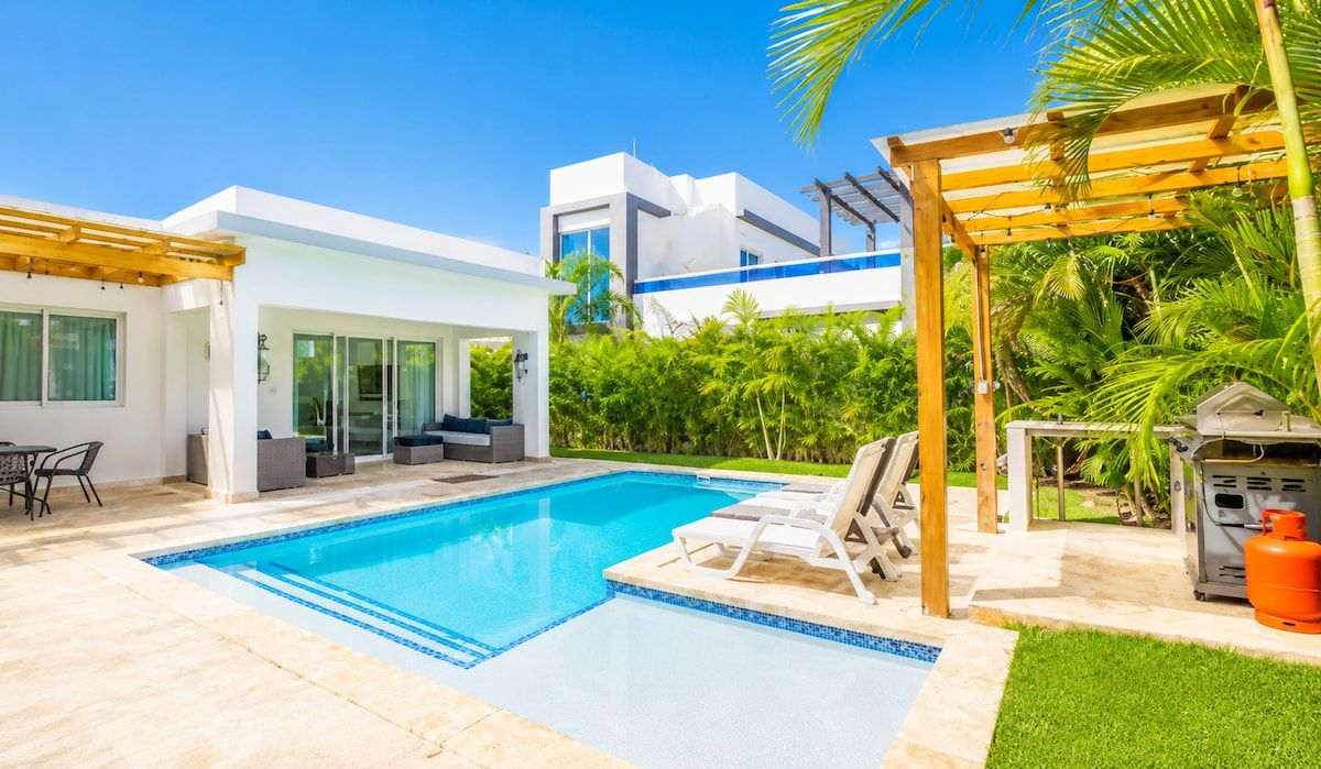 Ett modernt poolområde med en klarblå pool, en träpergola och en gasolgrill.