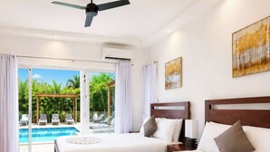 A hotel room with two beds, a ceiling fan, an air conditioning unit, a painting, and a view of a pool and palm trees. - Family Villa | Bunk Room, Pool & Up to 12 Guests (Sosúa)