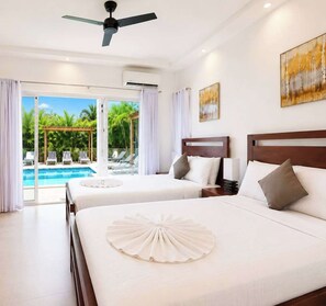 Ein Hotelzimmer mit zwei Betten, einem Deckenventilator, einer Klimaanlage, einem Gemälde und Blick auf einen Pool und Palmen.