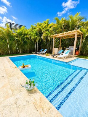 Ein klares, blaues Schwimmbecken mit Liegestühlen und Grillplatz, umgeben von üppigem Grün.