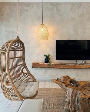 Ein modernes Wohnzimmer mit einem hängenden Rattan-Sessel, einer gewebten Pendelleuchte, einem Flachbildfernseher, einem Couchtisch aus Holz und einer rustikalen Holzregal.