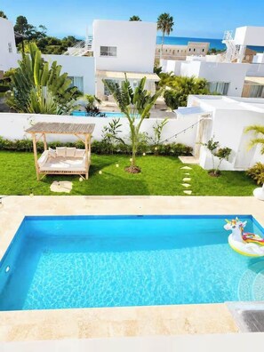 A swimming pool with a wooden deck and a lounge chair, surrounded by a well-maintained lawn and tropical plants. - Boho Retreat |Pool Cabana, Terrace Lounge, Rooftop (Sosúa)