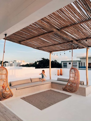 Ein Außenbereich mit Sitzgelegenheiten, einer Holz-Pergola, gepolsterten Bänken und hängenden Rattanschaukelstühlen.