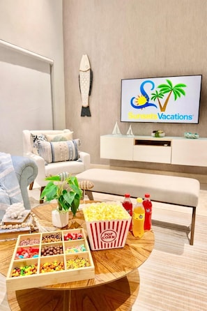 A modern living room with a sofa, a coffee table with snacks, a TV displaying a vacation ad, and a wall-mounted shelf. - Villa Caracol in Sosua Ocean Village Santa Fe (Sosúa)