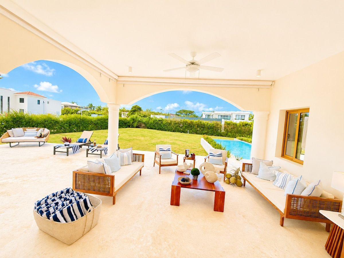A covered outdoor seating area with a pool view, furnished with sofas, chairs, and a coffee table.