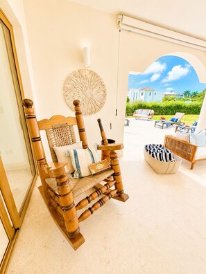 A wooden rocking chair with cushions and a woven basket, a wall-mounted decorative piece, and a view of a pool area with lounge chairs. - Sleeps up to 22 | Estate Pool 6bd Villa Corner Lot (Sosúa)