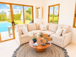 A modern living room with a beige sofa, a round wooden coffee table, and a view of a swimming pool. - Sleeps up to 22 | Estate Pool 6bd Villa Corner Lot (Sosúa)