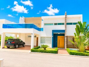 A modern two-story house with a car parked in the driveway, a wooden door, and a balcony with blue railings. - Sleeps up to 16 | Family Villa w/ Bunk Room & Spa (Sosúa)