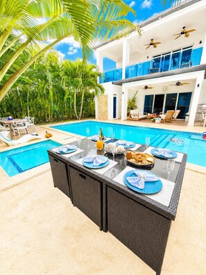 A poolside dining area with a table set for two, surrounded by lush greenery and a clear blue sky. - Sleeps up to 16 | Family Villa w/ Bunk Room & Spa (Sosúa)