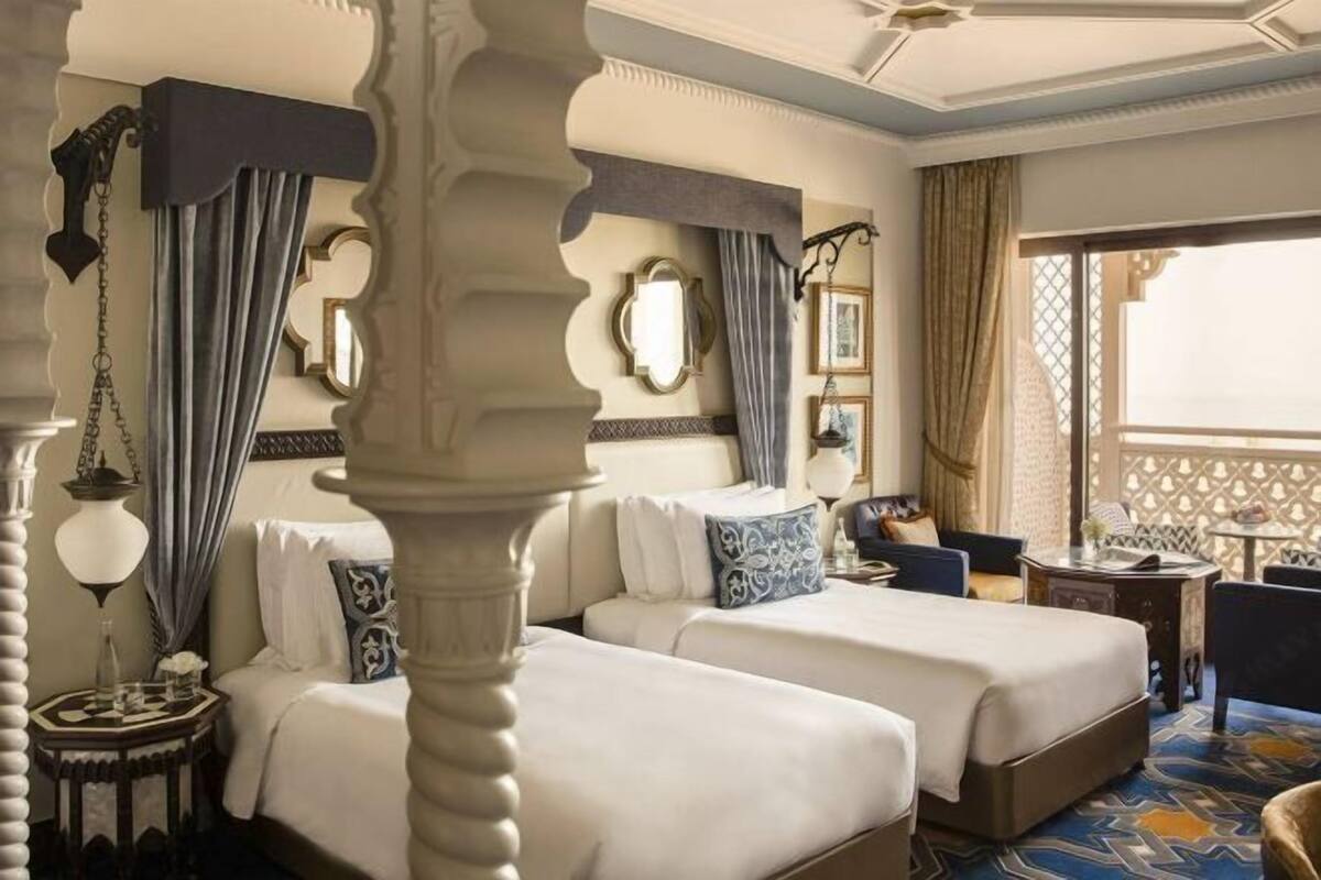 Habitación de hotel con dos camas, una mesita, una silla y un ventanal con vistas.