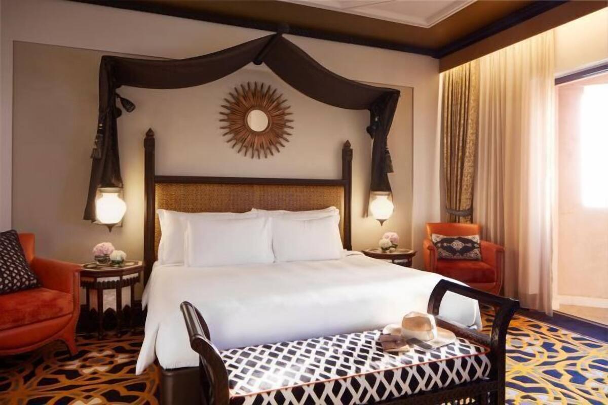 Una habitación de hotel con una cama grande, dos mesitas de noche, una silla, un espejo decorativo con diseño de sol y una alfombra estampada.