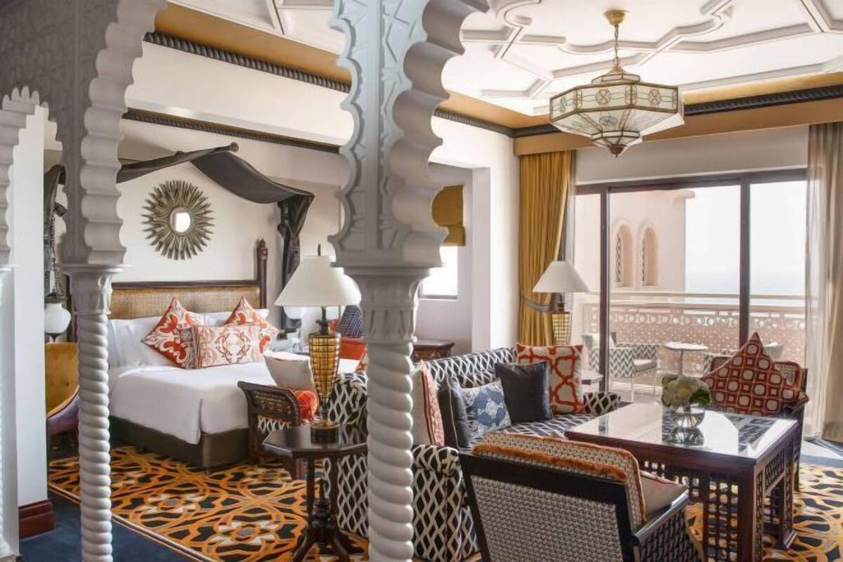 Habitación de hotel con cama, sofá, mesa de comedor y balcón con vistas.