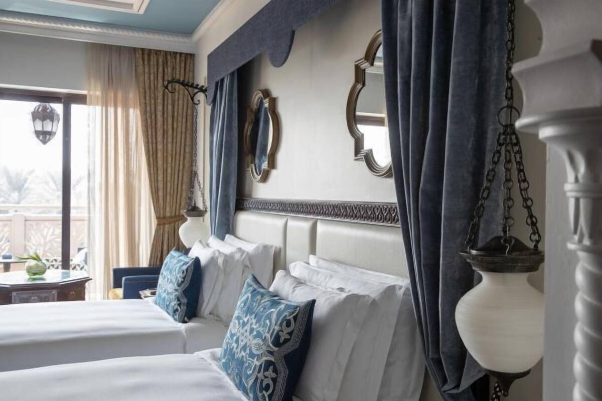 Habitación de hotel con dos camas, un ventanal con cortinas, una lámpara colgante y un espejo decorativo.