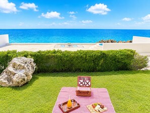 A picnic setup with a checkered blanket, a wicker basket, and a tray with food and drinks on a grassy area with a view of the ocean. - Oceanfront Paradise | Cliffside Spa & Pool in SOV (Sosúa)