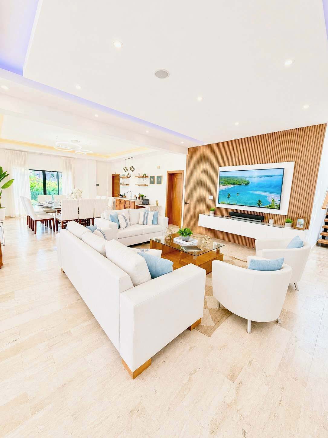 A modern living room with a large flat-screen TV mounted on a wooden wall, a dining area with a table and chairs, and a kitchen in the background.
