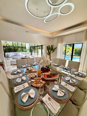 A dining area with round tables, white chairs, and a large window with a view of greenery and a pool. - 8 BR Grand Villa in Oceanfront Comm Sosua Ocean Vill (Sosúa)