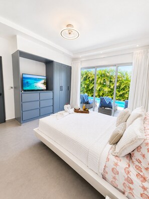 A bedroom with a large bed, a television on a built-in shelf, and a view of a pool and greenery. - Sunset Villa | Bunk Room, Pool & Sleeps up to 11 (Sosúa)