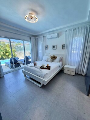 A modern bedroom with a large bed, a bedside table, a small bedside cabinet, a ceiling light, and a view of the outdoors. - Sunset Villa in Oceanfront Sosua Ocean Village (Sosúa)