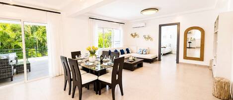 A spacious living area with a dining table set for four, a sofa, and a large window offering a view of greenery.