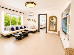 Een moderne woonkamer met een bank, salontafel en televisie. Er is een groot raam met gordijnen, een spiegel en een deur die naar een andere kamer leidt.