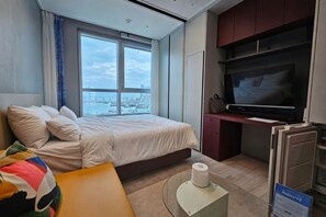 A hotel room with a bed, a desk, a television, and a window with a city view. - The Bayment Rasidence (Busan)