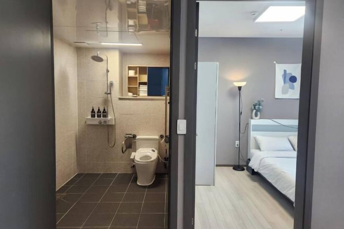 A bathroom with a shower, toilet, and a bed in the adjacent room.