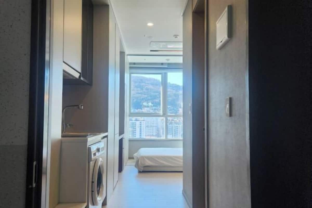 A narrow hallway leading to a bedroom with a large window, a washing machine, and a bed.