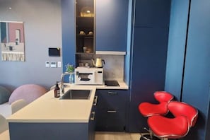 A compact kitchen with a microwave, sink, and cabinets, a small seating area with a chair, and a wall-mounted shelf with decorative items. - The Bayment Rasidence (Busan)