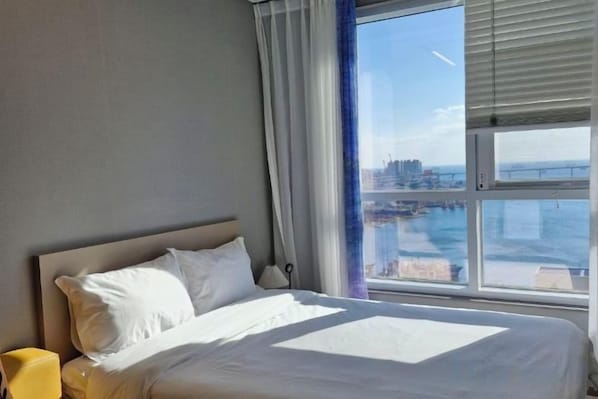 A hotel room with a large bed, a nightstand, and a view of a cityscape through a window. - The Bayment Rasidence (Busan)