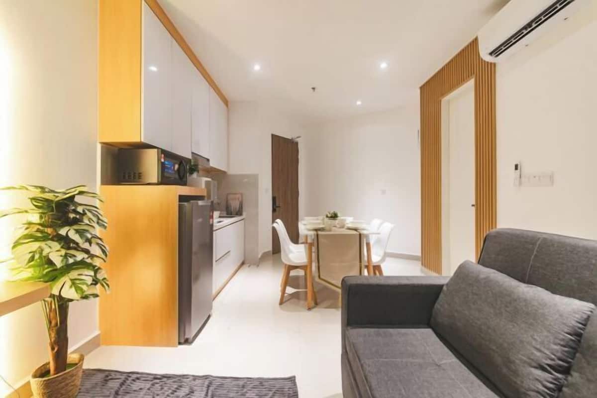 A modern living room with a grey sofa, a dining area with white chairs, and a kitchen with wooden cabinets.