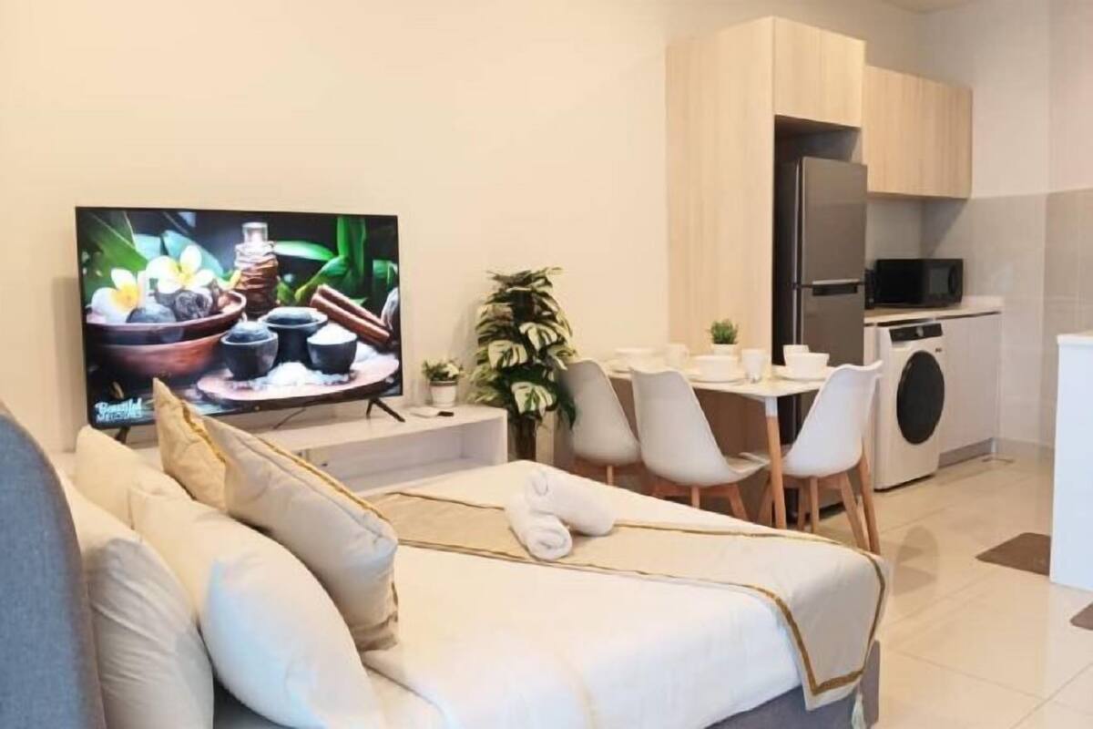A modern hotel room with a sofa, a TV, a dining area with chairs, a kitchen with a microwave, and a washing machine.