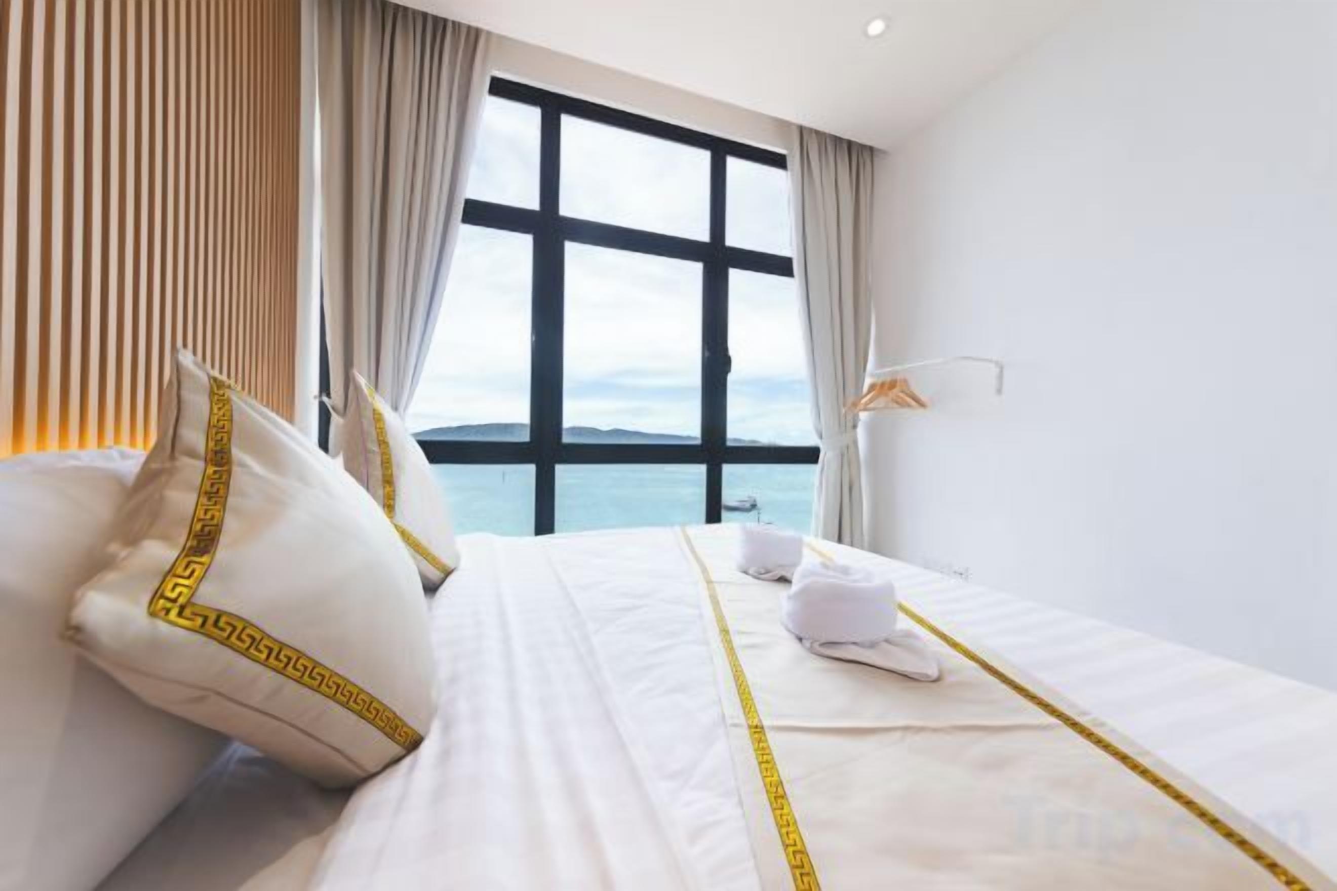 Habitación de hotel con un ventanal grande, una cama con ropa blanca y almohadas decorativas, y vista al mar.