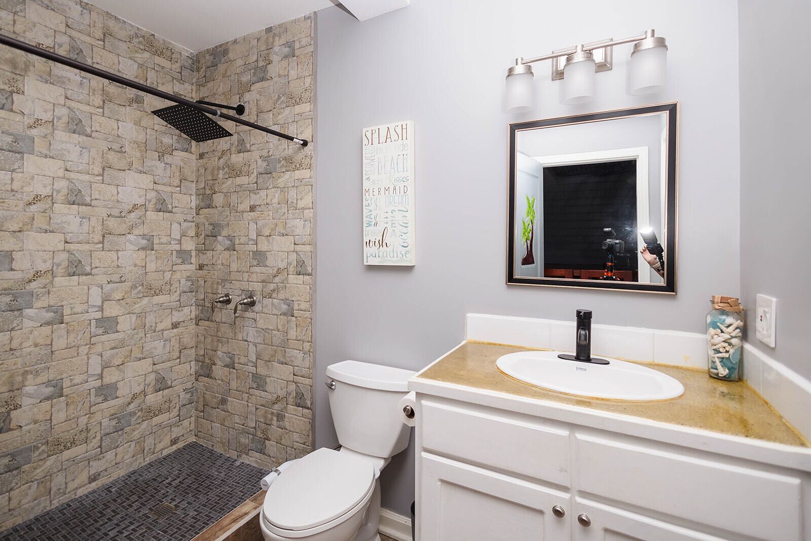 Un baño con pared de ducha de piedra, inodoro blanco, lavabo con espejo y un cuadro enmarcado en la pared.