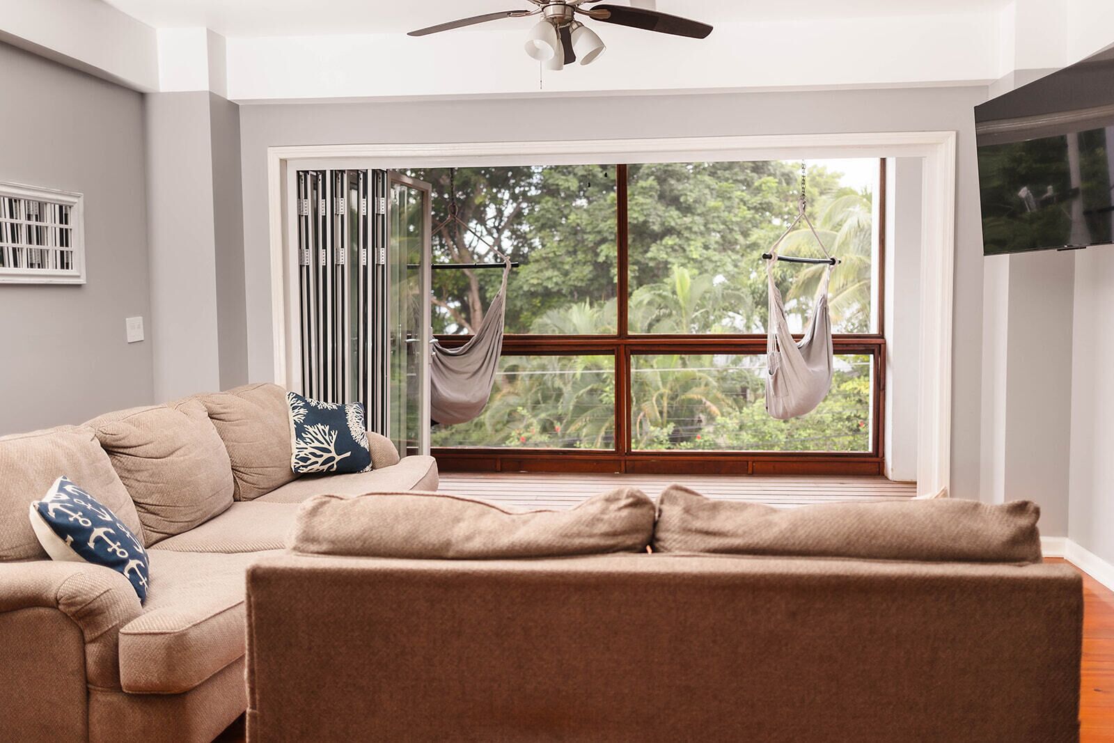 Un salón con un sofá color crema, un ventilador de techo y un ventanal con vistas a la vegetación.
