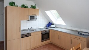 Eine Küche mit Holzausstattung, einem Einbauofen, einer Mikrowelle und einem Spülbecken. Über dem Spülbecken befindet sich ein Oberlicht und auf der rechten Seite ein kleines Fenster.