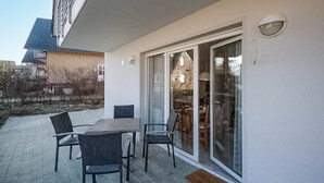 Ein überdachter Außenbereich mit Tisch und Stühlen, eine Schiebetür zu einem Raum und ein Gebäude im Hintergrund.