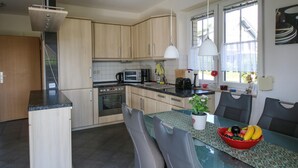 Een moderne keuken met lichte houten kastjes, een ingebouwde oven, een magnetron en een koffiezetapparaat. Er staat een eettafel met een rode fruitschaal en een potplant.