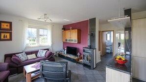 Een moderne woonkamer met een bank, televisie en open haard. Er is een keuken met een fruitschaal en een eethoek met een tafel en stoelen.