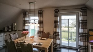 Un coin repas avec une table en bois, des chaises, un vase de fleurs et une vue sur l’extérieur grâce à de grandes fenêtres ornées de rideaux à carreaux.