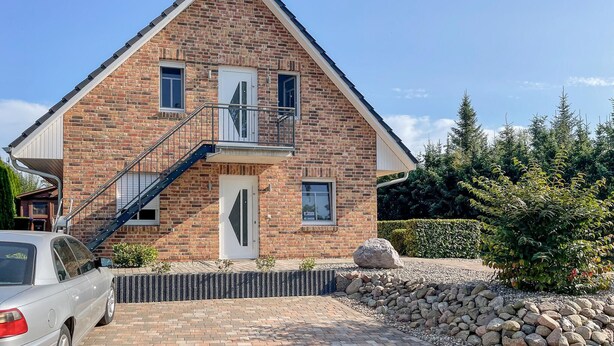 Une maison à deux étages, construite en briques, avec une entrée à l’avant, un petit balcon et une voiture garée devant.