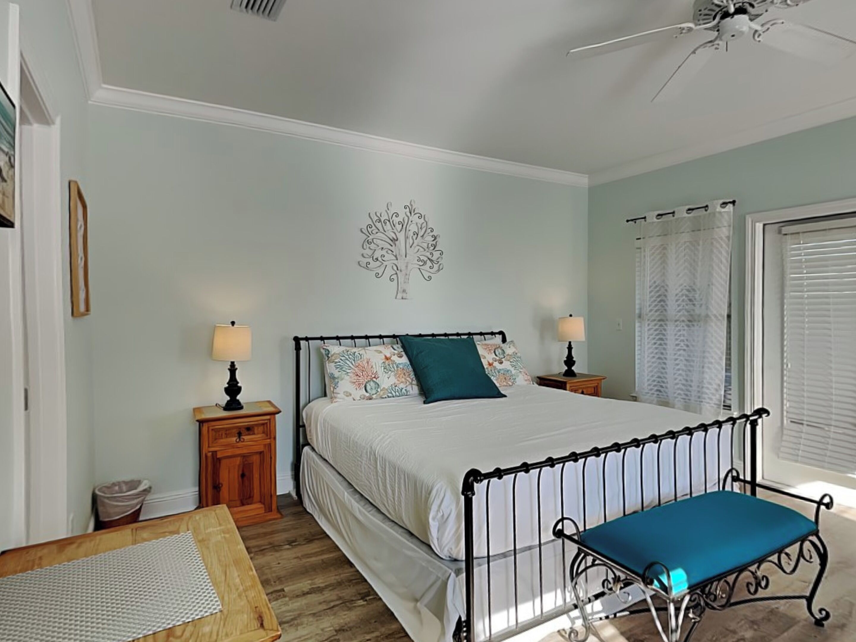 A bedroom with a bed, two bedside tables, a nightstand, a wooden bench, a ceiling fan, and a wall-mounted tree decoration.
