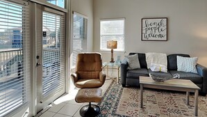 Een woonkamer met een bruine leren fauteuil, een grijze bank, een bijzettafeltje en een vloerkleed. Er is een raam met zonwering, een lamp en een bord op de muur.