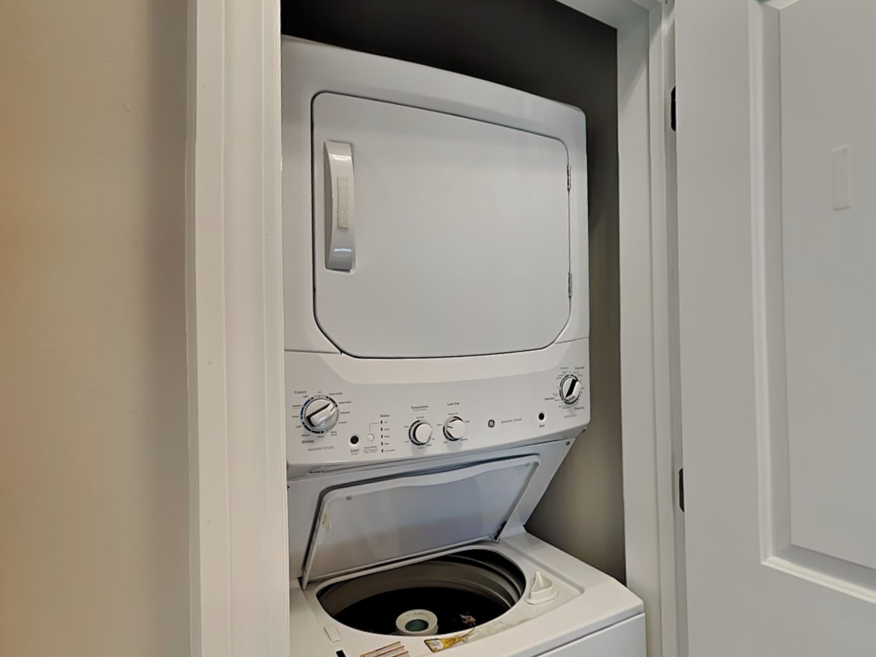 A compact, white front-loading washer and dryer unit with control knobs and a transparent door, installed in a small laundry room with a plain wall and a door visible to the right.