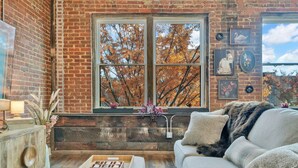 Ein gemütliches Zimmer mit Backsteinwand, einem großen Fenster mit Herbstlaub, einem weißen Sofa und einem Holznachttisch mit Lampe.