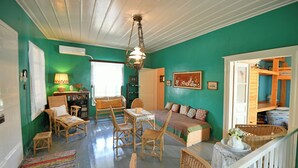 A room with teal walls, a wooden ceiling, wicker furniture, a bed with a green and white patterned bedspread, and a white railing. - Villa Ariadne - Sea View Sanctuary (Idra)