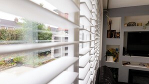 A living room with a black leather sofa, a flat-screen TV on a white shelf, and a view of a garden through white blinds. - Beautiful Wythenshawe Home Near Airport With Garden & Parking Sleeps 5 (Manchester)