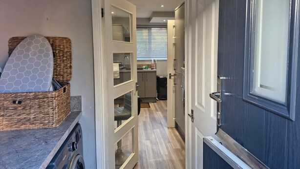 A narrow hallway leading to a kitchen with a washing machine, a basket with a surfboard, and a door with a window. - Beautiful Wythenshawe Home Near Airport With Garden & Parking Sleeps 5 (Manchester)