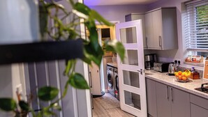 A modern kitchen with grey cabinets, a washing machine, and a countertop with fruits and a coffee maker. - Beautiful Wythenshawe Home Near Airport With Garden & Parking Sleeps 5 (Manchester)